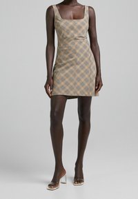 Ljusbrun rutmönstrad klänning med fyrkantig halsringning, figurnära passform och minilängd. Bärs med genomskinliga klacksandaler.