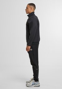 Mann in schwarzer Zip-Jacke und passenden Jogginghosen mit weißen und blauen Laufschuhen, der seitlich vor einem schlichten Hintergrund steht.