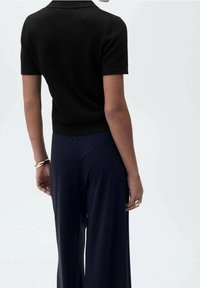 Polo noir en maille avec manches courtes, taille ajustée, associé à un pantalon large bleu marine, bijoux dorés visibles au poignet.