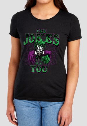 Woman wearing black t-shirt featuring Joker with green hair, purple suit, and text "The Joker's on you" in green and purple lettering.