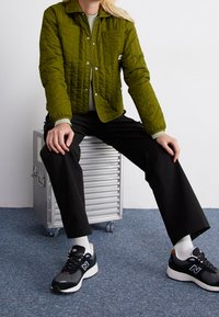 Olivgrön quiltad jacka med tryckknappar och två bröstfickor; kombinerad med svarta byxor och svarta New Balance-sneakers.