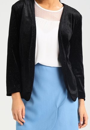 Vrouw draagt een doorschijnend wit blouse met een zwarte fluwelen blazer met open voorkant en een lichtblauwe rok met hoge taille, staand tegen een effen achtergrond.