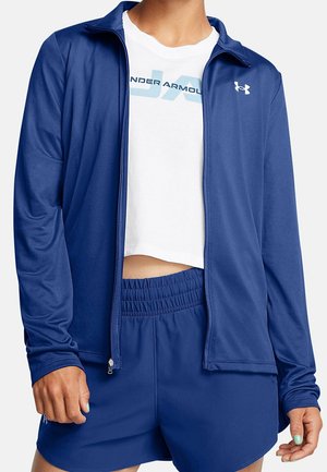 Person iført en hvid Under Armour crop top, blå lynlåstrøje og matchende blå atletiske shorts med elastisk talje, stående med afslappede hænder.