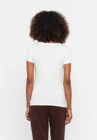 T-shirt branco de manga curta com decote arredondado, tecido suave e um corte descontraído. Usado com calças castanhas, exibindo um design simples.