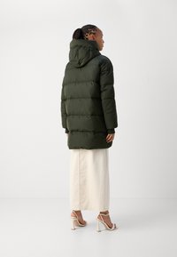 Donkergroen gewatteerd parka-jack met een capuchon, voorzien van horizontale doorgestikte naden en geribbelde manchetten, gedragen over een lichtcrème rok en beige hakken.