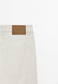 Vue arrière d’un jean en denim beige clair avec une seule poche arrière et un patch en cuir marron portant le nom de la marque Massimo Dutti.