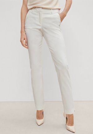Nainen, jolla on päällään off-white väriset slim-fit housut ja yhteen sopivat korkokengät, seisoo yhdellä kädellä taskussa yksiväristä taustaa vasten.