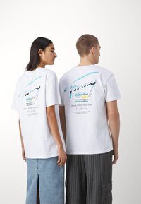 Dos personas de pie, espalda con espalda, vestidas con camisetas gráficas blancas con el logo y texto "Tommy", combinadas con una falda de mezclilla y pantalones de rayas.