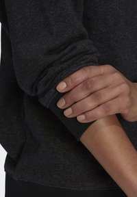 Czarna bluza z miękkiej, teksturowanej tkaniny. Mankiet wyposażony w dopasowaną, elastyczną opaskę. Dłoń spoczywa na rękawie, ukazując gładką skórę.