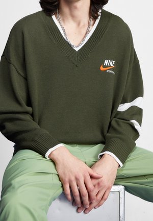 Person trägt olivgrünen Nike-Pullover mit weißen Streifen an den Ärmeln und hellgrüne Hose, Hände ruhen auf den Knien im Sitzen.