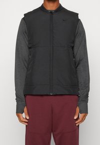 Schwarze ärmellose Steppweste mit einem Frontreißverschluss, hohem Kragen und einem kleinen Logo auf der linken Brust. Darunter wird ein graues Langarmshirt getragen.