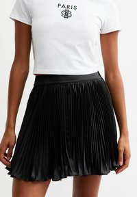 Falda negra con pliegues y un acabado suave de satén, con una cinturilla ancha, combinada con una camiseta corta blanca con texto bordado en negro.