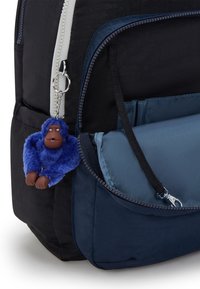 Zwarte stoffen rugzak met een blauwe nep bont aap sleutelhanger. Voorzien van een voorvak met rits en een grote zijcompartiment met een contrasterend blauw paneel.