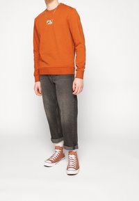 Persona con sudadera naranja con un logo blanco, jeans oscuros con puños, calcetines blancos y zapatillas altas naranjas, de pie sobre un suelo blanco.