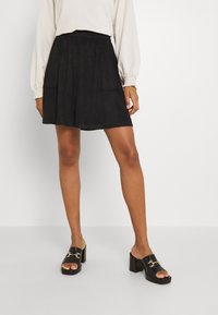 Czarna spódnica zamszowa z elastycznym paskiem w talii, z dwoma przednimi kieszeniami. Zestawiona z czarnymi sandałami na platformie typu slip-on, ozdobionymi złotymi elementami.