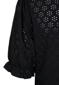 Blouse noire en tissu texturé et perforé avec un motif floral. Les manches bouffantes se terminent par un détail de poignets volantés.