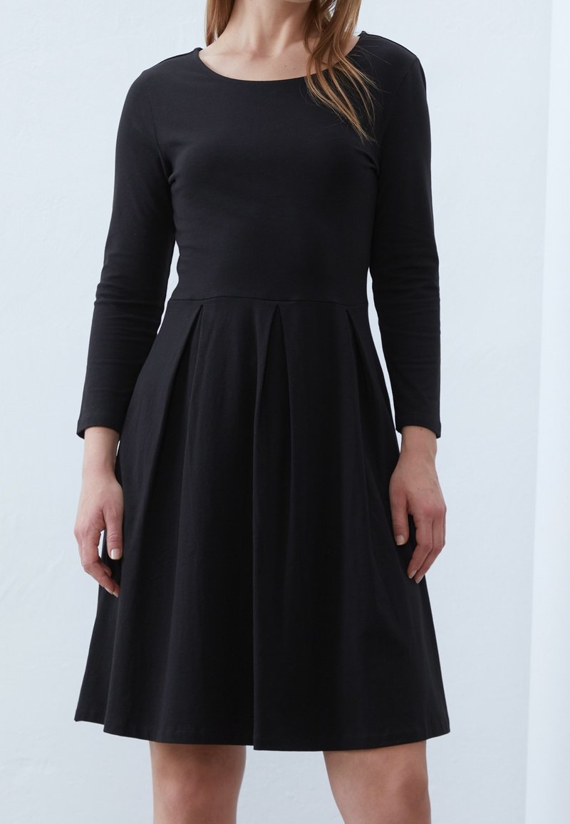 Femme portant une robe noire à manches longues, longueur genou, avec un corsage ajusté et une jupe plissée, debout devant un fond clair.