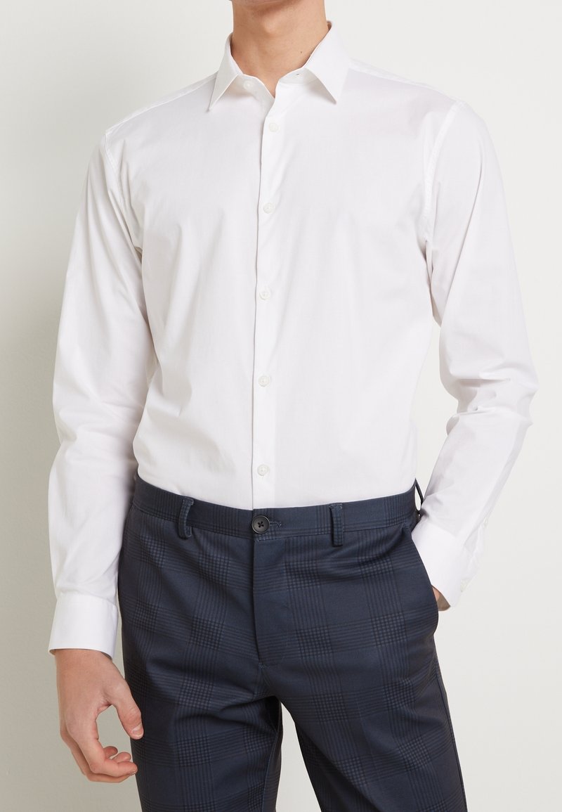 Homme portant une chemise blanche ajustée à manches longues boutonnée, rentrée dans un pantalon à carreaux bleu foncé, avec une main dans la poche.