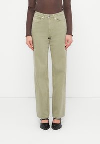 Άτομο που φοράει ελιάς ίσια παντελόνια, καφέ μακρυμάνικη μπλούζα και μαύρα μυτερά παπούτσια Mary Jane, στέκεται μπροστά σε ένα λευκό φόντο.