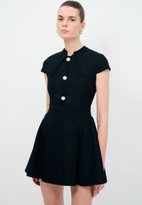 Schwarzes Kleid mit tailliertem Oberteil, kurzen Ärmeln und schwingendem Rock. Verfügt über eine Spitzenstruktur, einen Schleifenkragen und drei dekorative Knöpfe.