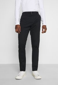 Chinos de algodón negros con un diseño ajustado, que cuentan con un cierre de botón en la parte delantera, bolsillos laterales y una textura suave. Combinados con zapatillas blancas.