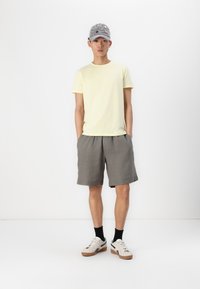 Lichtgele korte mouwen shirt, grijze shorts en casual sneakers met zwarte accenten. Grijze pet met decoratieve elementen; staande pose.