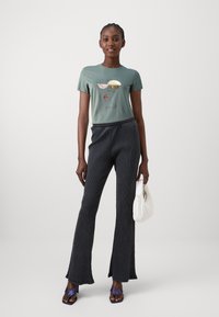 T-shirt en coton vert avec design graphique, associé à un pantalon évasé en côte sombre. Accessoirisé avec un sac à main matelassé blanc et des sandales violettes.