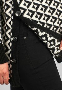 Sweter z dzianiny z czarno-białym wzorem geometrycznym, zapinany na guziki, z ściągaczami przy mankietach. Połączony z dopasowanymi czarnymi spodniami.