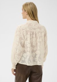 Femme aux longs cheveux blonds portant un chemisier transparent crème à motif floral avec col à volants et poignets froncés, associé à un pantalon en cuir marron foncé.