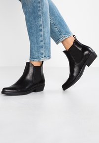 Bottines en cuir noir avec des panneaux élastiques sur les côtés et un petit talon carré. La texture est lisse avec un léger éclat.
