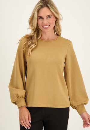 Lachende vrouw met lang blond haar draagt een mosterdgele top met lange mouwen en blousende mouwen en een zwarte broek, staand tegen een effen achtergrond.