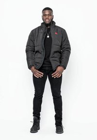 Svart pufferjacka med hög krage, dragkedjestängning och röd logodetalj; bärs över en svart skjorta och matchas med svarta jeans och stövlar.