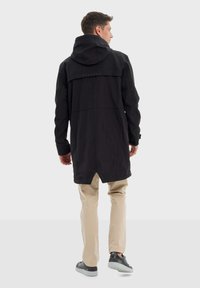 Manteau noir long avec capuche, présentant un col montant et des détails de couture subtils, associé à un pantalon beige neutre et des sneakers noires.