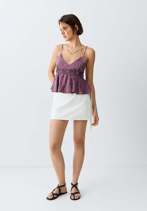 Jeune femme debout portant un top sans manches violet à motifs, une mini-jupe blanche, des sandales noires à lanières, ainsi qu'un collier simple et des boucles d'oreilles.