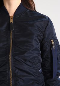 Giacca bomber della Marina realizzata in nylon lucido, con zip dorata, colletto a coste e tasca con zip impreziosita da dettagli in tessuto.