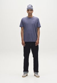 Vijolična bombažna majica s kratkimi rokavi, ki ima majhen logotip. Temno jeans hlače in svetlo siva pletena kapa dopolnjujeta outfitt.