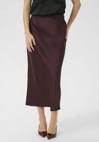 Burgundy satin maxikjol med en slät textur och en figurnära midja, parat med glänsande spetsiga klackar och en svart topp med spetsdetaljer.