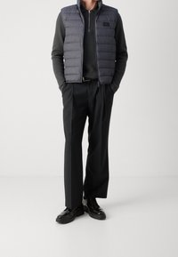 Szara, pikowana kamizelka na ciemnej koszuli z długim rękawem, w połączeniu z czarnymi spodniami i czarnymi butami. Kamizelka ma wysoki kołnierz i zamki błyskawiczne.