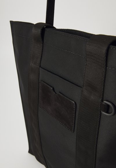 Black tote bag made of durable fabric. Features sturdy straps, a front pocket with a suede accent, and a sleek, simple design.