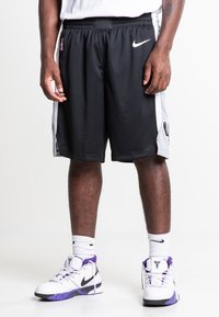 Zwarte basketbalshorts met witte zijpanelen, voorzien van een NBA-logo en een Nike swoosh. Draag met witte en paarse Nike-schoenen en -sokken.
