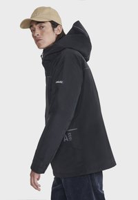 Veste imperméable noire avec capuchon, fermeture éclair et accents de logo. Associée à une casquette beige. Le mannequin pose de profil.