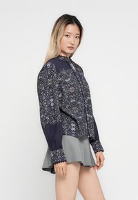 Young woman with long ombre hair wearing a dark floral long-sleeve blouse and grey pleated skirt, standing against a white background.