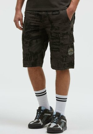 Mann trägt schwarze gemusterte Cargo-Shorts, weiße Crew-Socken mit schwarzen Streifen und schwarze und graue Sportschuhe, steht mit den Händen in den Taschen.