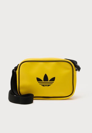 Rumena pravokotna torbica čez ramo s črnim logotipom Adidas trefoil in nastavljivim črnim trakom na belem ozadju.