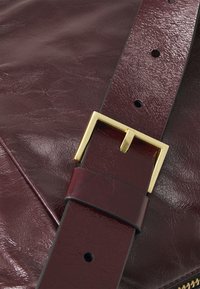 Dettaglio di una borsa in ecopelle bordeaux con una fibbia in metallo dorato su una tracolla. La texture è liscia con una leggera lucentezza e presenta cuciture visibili.