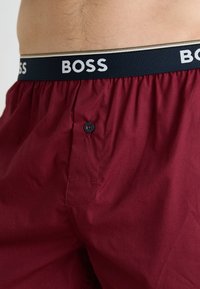 Bordeaux férfi bokszernadrág, elülső gombbal és fekete rugalmas derékpánttal, amelyen fehér szöveggel szerepel a "BOSS" márkajelzés.