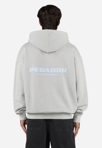 Lys grå hættetrøje med oversized pasform, ribbede manchetter og kant. Ryggen har stort blåt tekst: "PEGADOR" og "CERTIFICERET AUTENTISK GADEMODE".