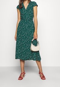 Grünes blumiges Wickelkleid mit kurzen Ärmeln, knielang, kombiniert mit einer kleinen beigen Handtasche und roten Riemchen-Heels.
