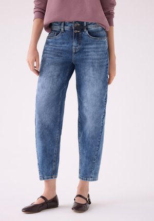 Femme portant un jean bleu délavé taille haute avec une fermeture à double bouton et des chaussures plates marron foncé, debout devant un fond uni.