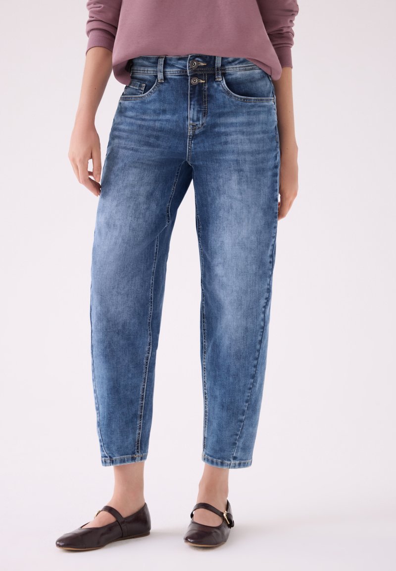 Femme portant un jean bleu délavé taille haute avec une fermeture à double bouton et des chaussures plates marron foncé, debout devant un fond uni.